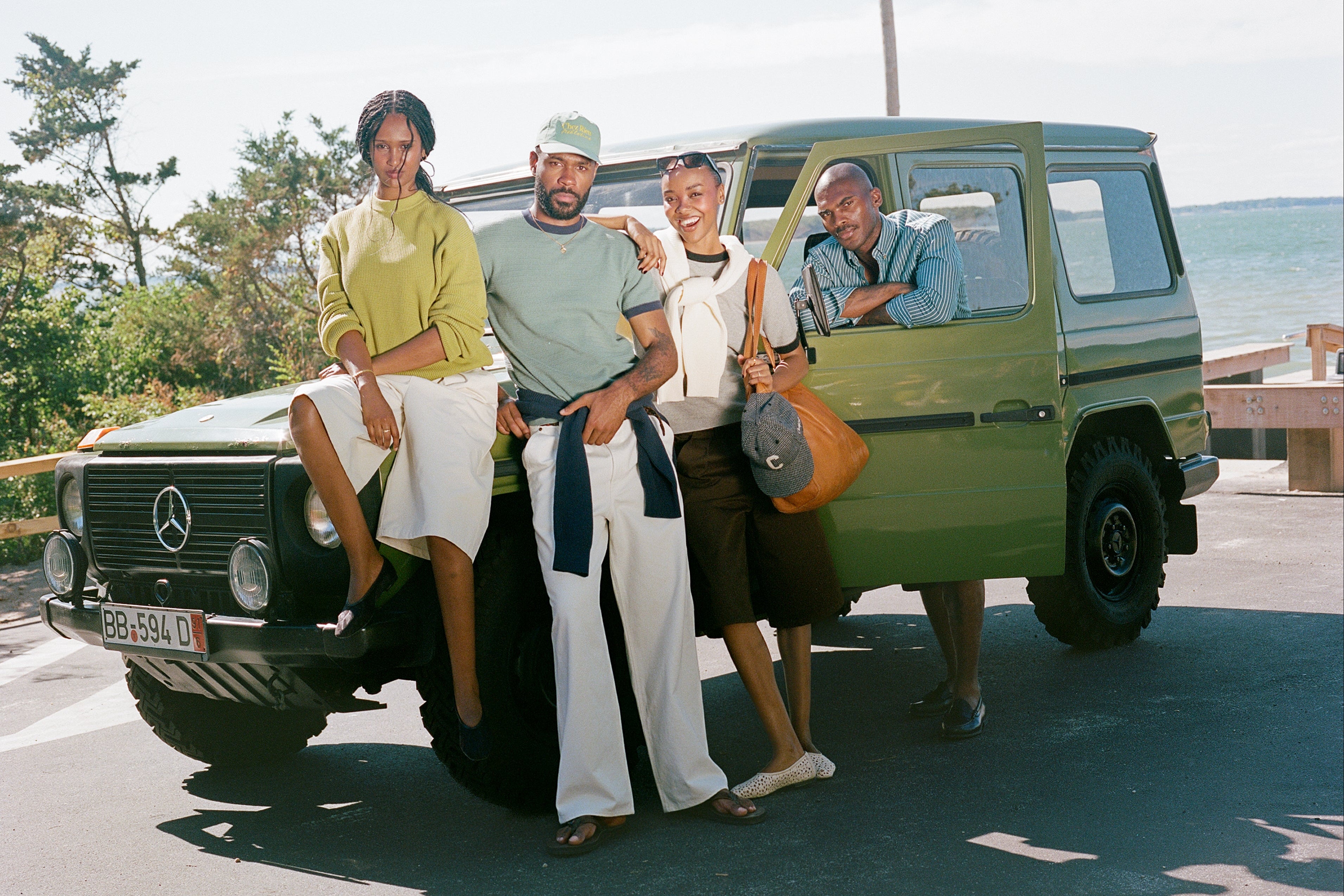 Four people posing with a vintage olive green Mercedes-Benz G-Class SUV on a paved road with water and trees in the background.